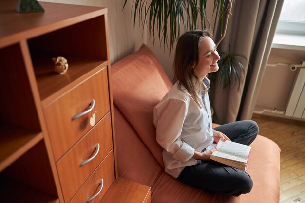 Mulher jovem, alegre, confiante e feliz relaxando em sua poltrona aconchegante em casa com livros